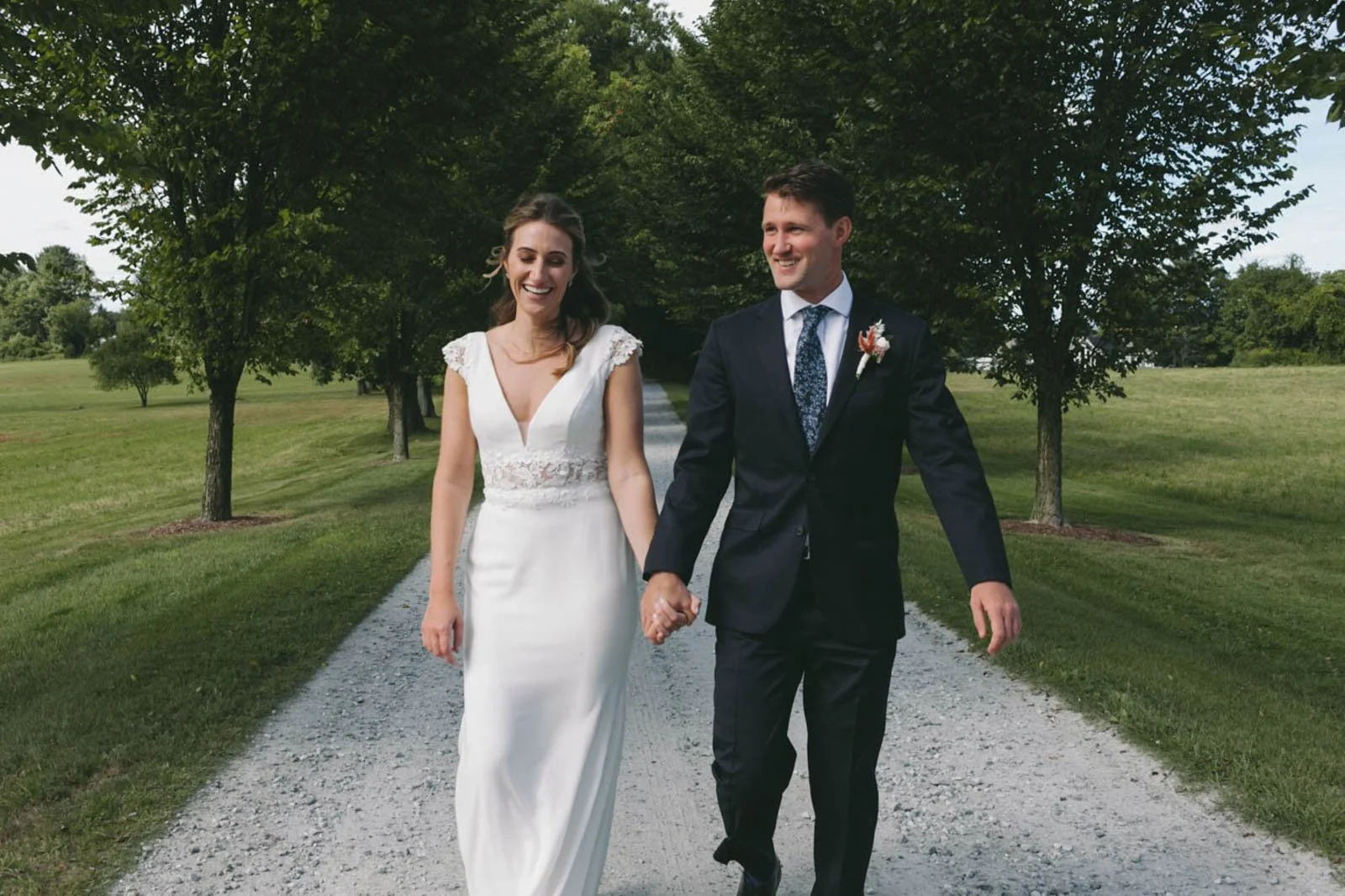A newlywed couple walking hand-in-hand outdoors on a gravel path surrounded by green grass and trees, smiling happily.