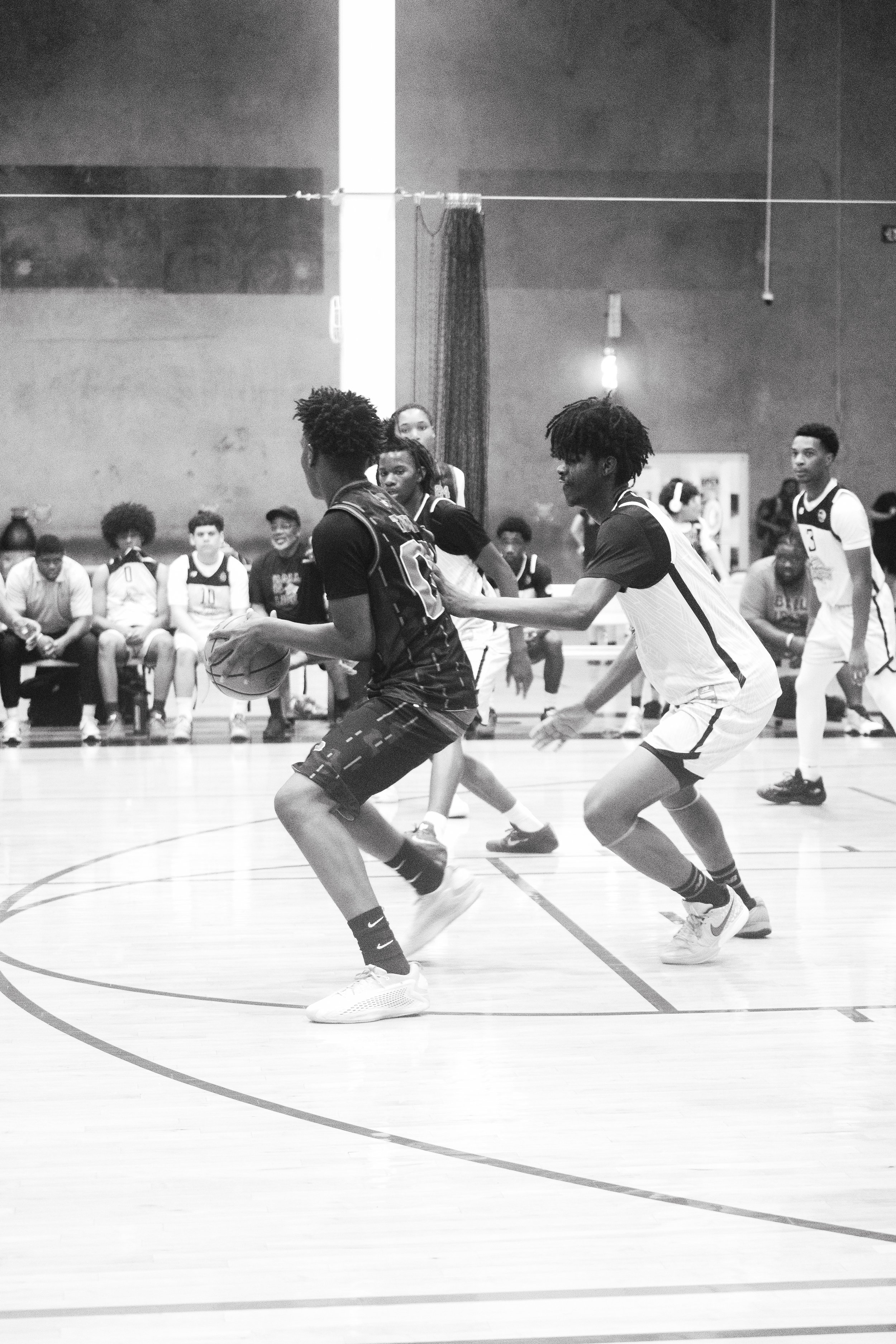 Black and white photo of a basketball game showing players in action on the court with spectators sitting on the sidelines.