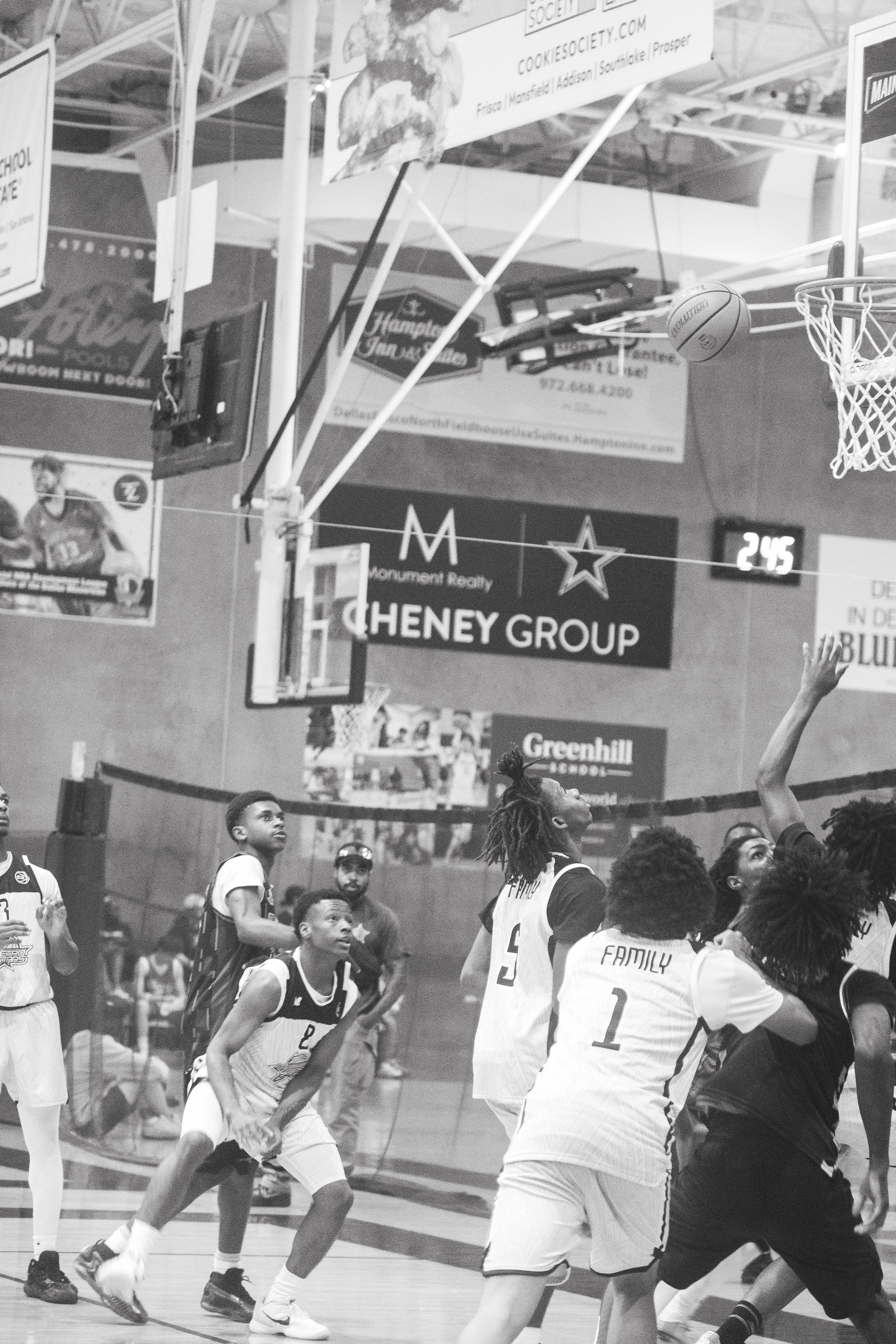 Basketball game with players on the court, some watching and others reaching for the ball near the hoop; basketball in mid-air near the goal; gymnasium with advertisements and people in the background.