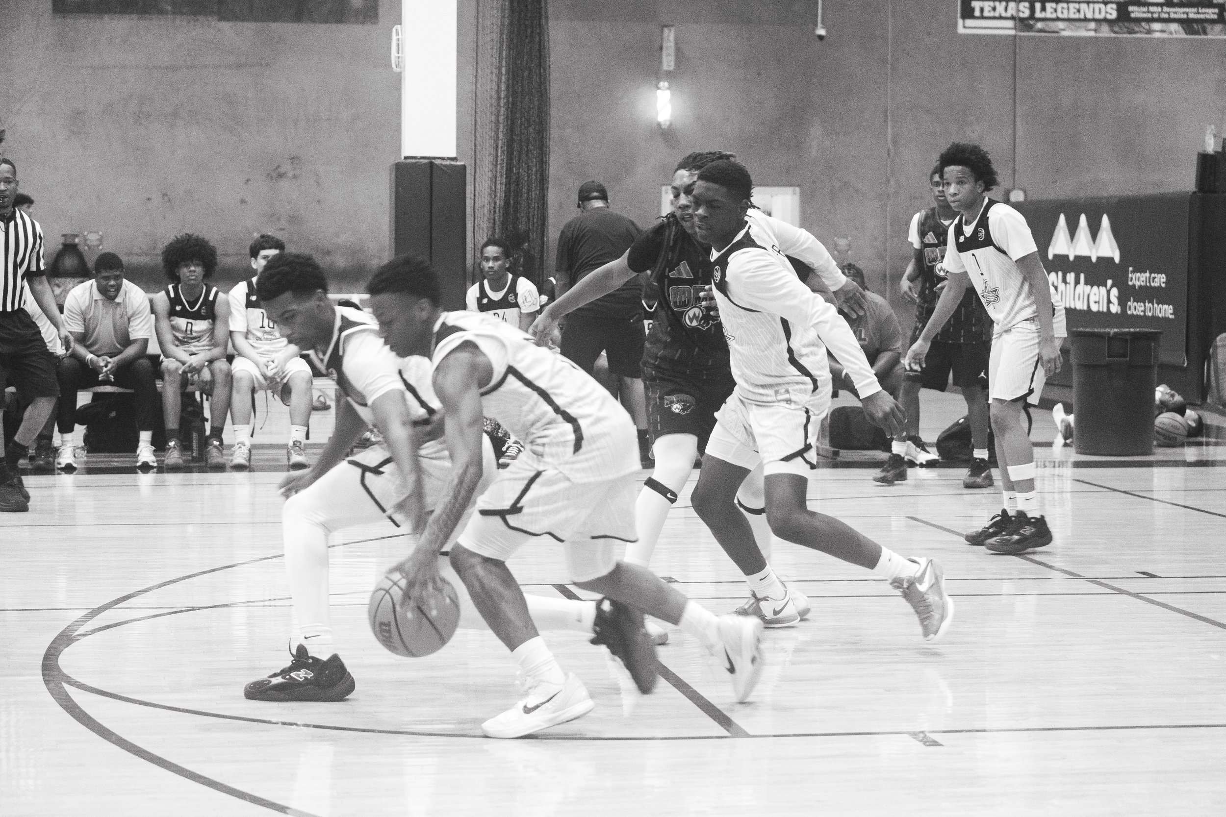 A black and white photo of a youth basketball game where players are competing for the ball on the court. Some players are sitting on the bench watching, and a referee is present on the left side of the image. The scene is inside a gym with banners a