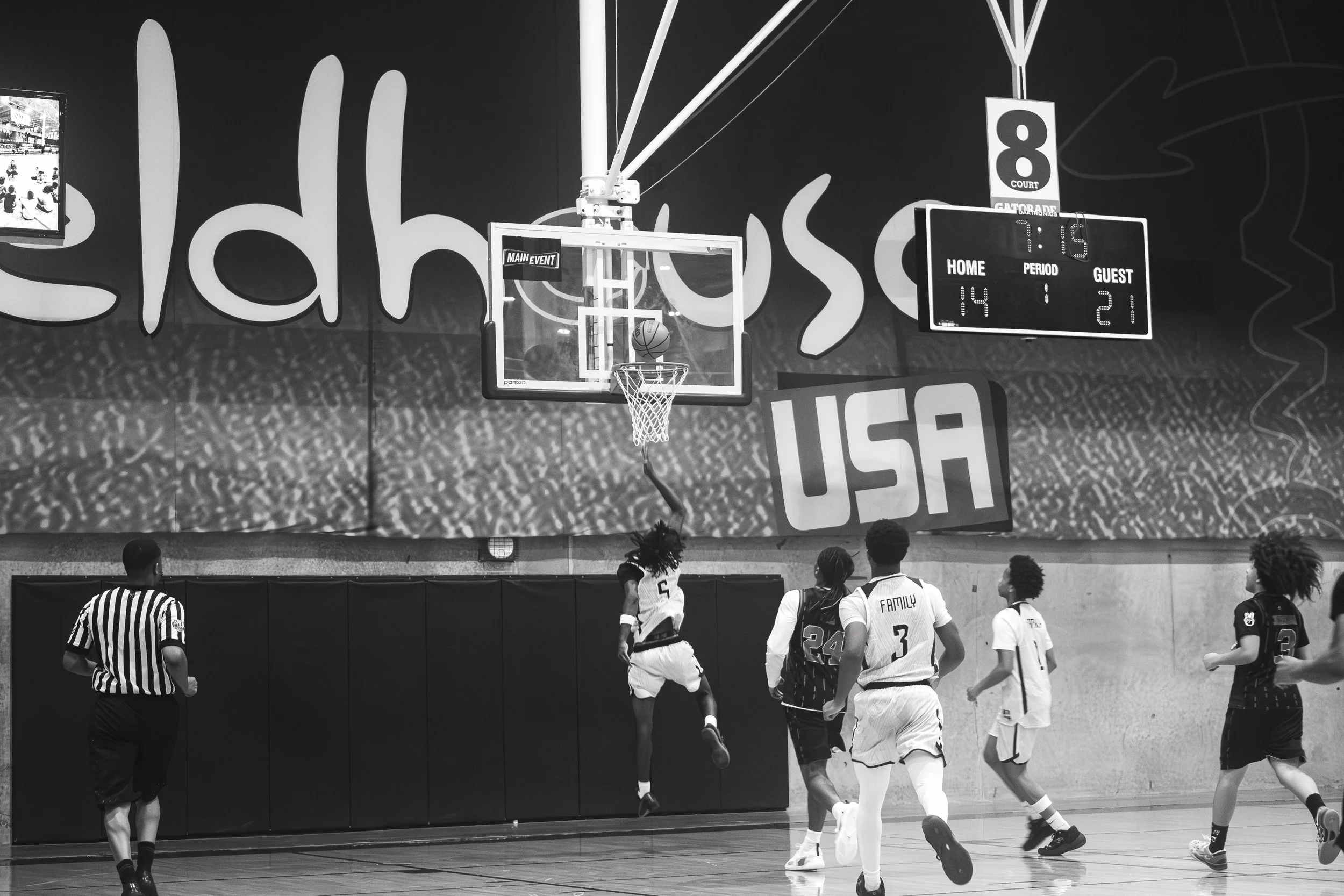 A basketball game is in progress with players on the court, a scoreboard showing scores of 14-21 with 1:45 remaining in the period, and a basketball hoop visible in the background. A referee is standing near the players, and there is a large sign wit