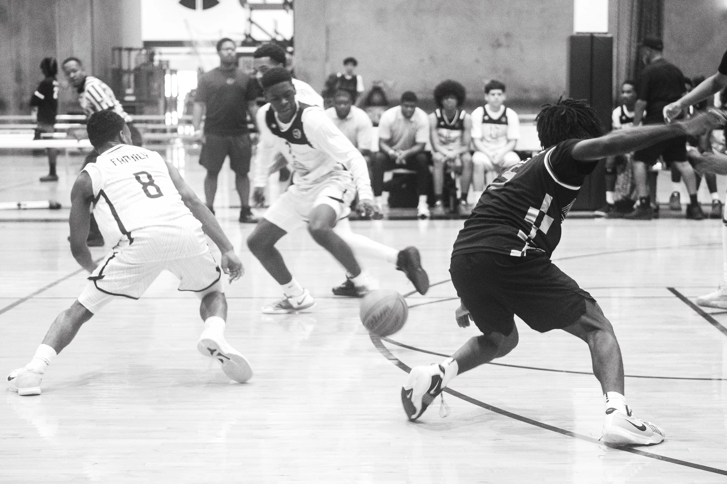 Young men playing basketball on an indoor court with spectators and officials watching in the background.