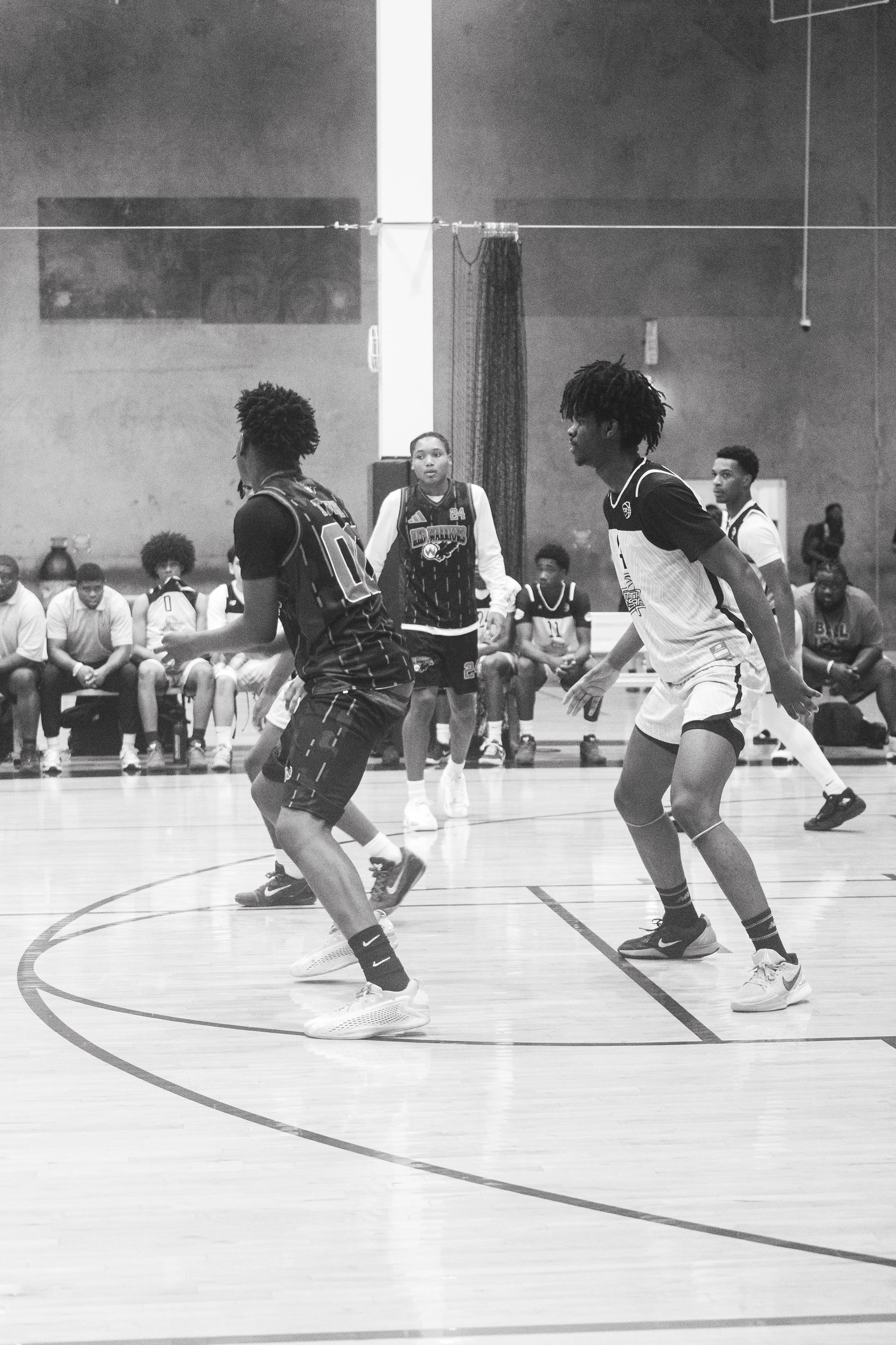Four basketball players in action on an indoor court, with spectators seated on the sidelines, some wearing jerseys and sitting in chairs; indoor gym setting with a high ceiling.