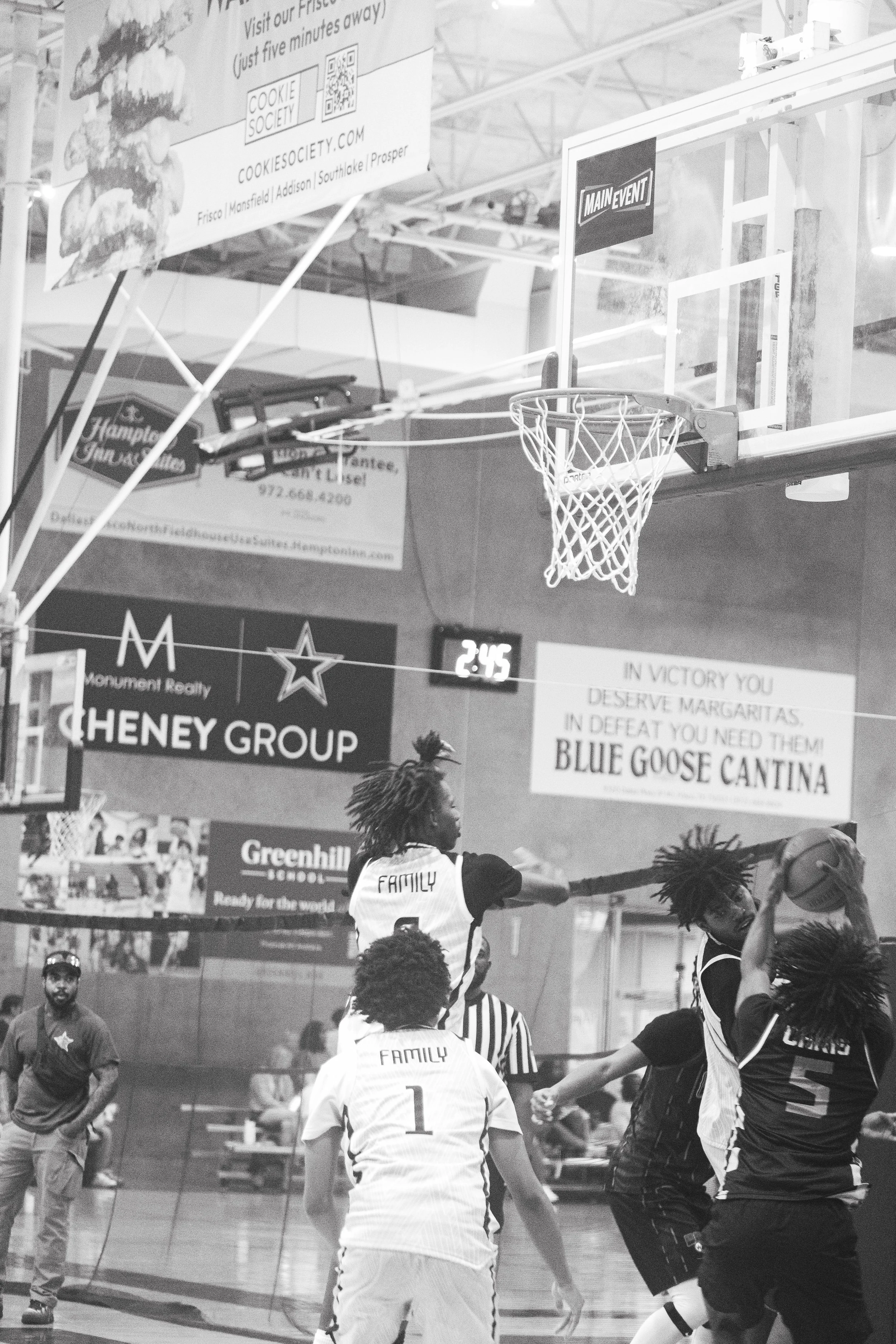 Basketball game with players reaching for the ball near the hoop, referee on the court, spectators in the background, and advertising banners hanging from the ceiling.
