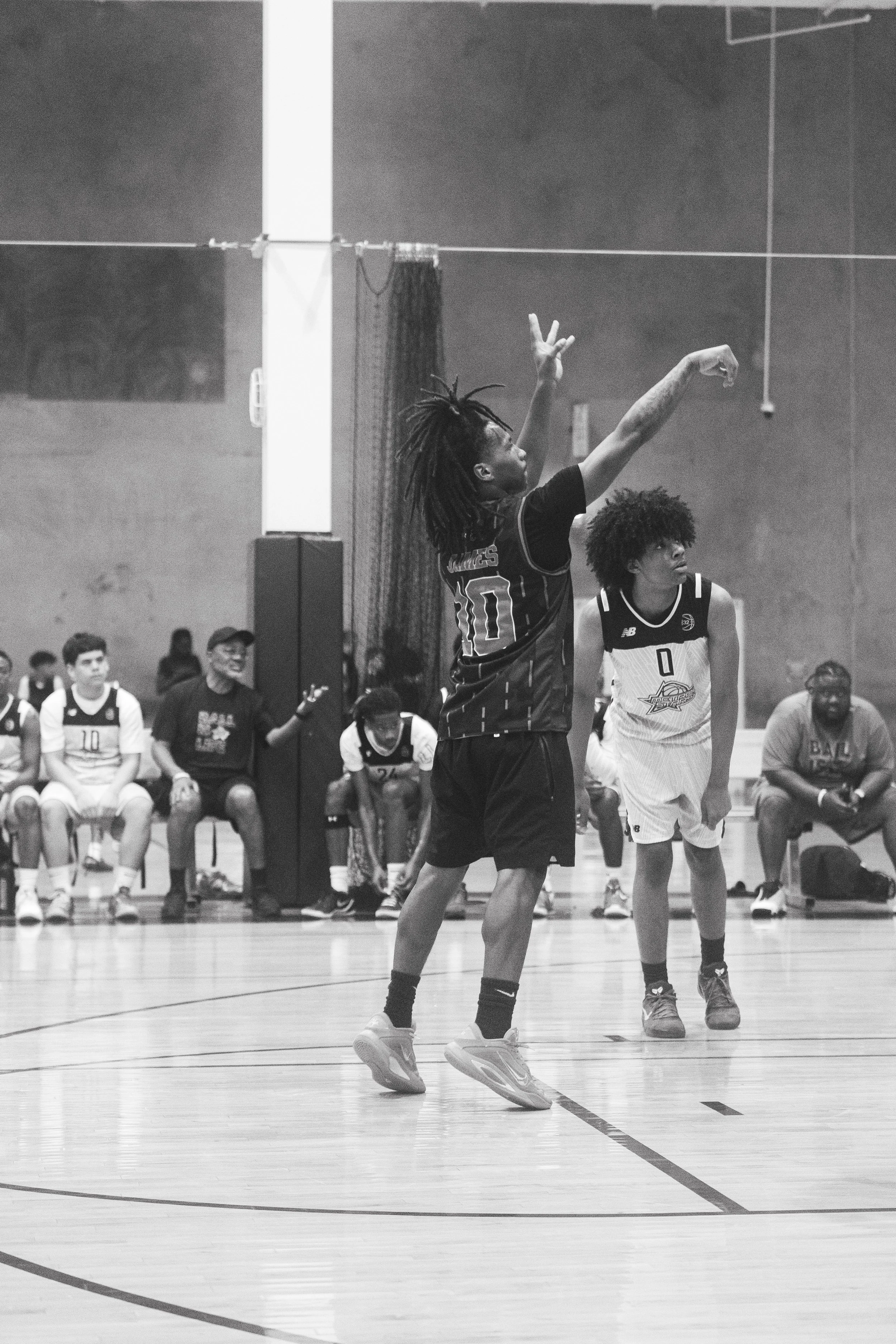 A black-and-white photo of a basketball game in progress. Two players are in focus, with one preparing to shoot or pass the ball in mid-air. The player in the foreground has dreadlocks and is wearing a dark jersey, while the other player has curly ha