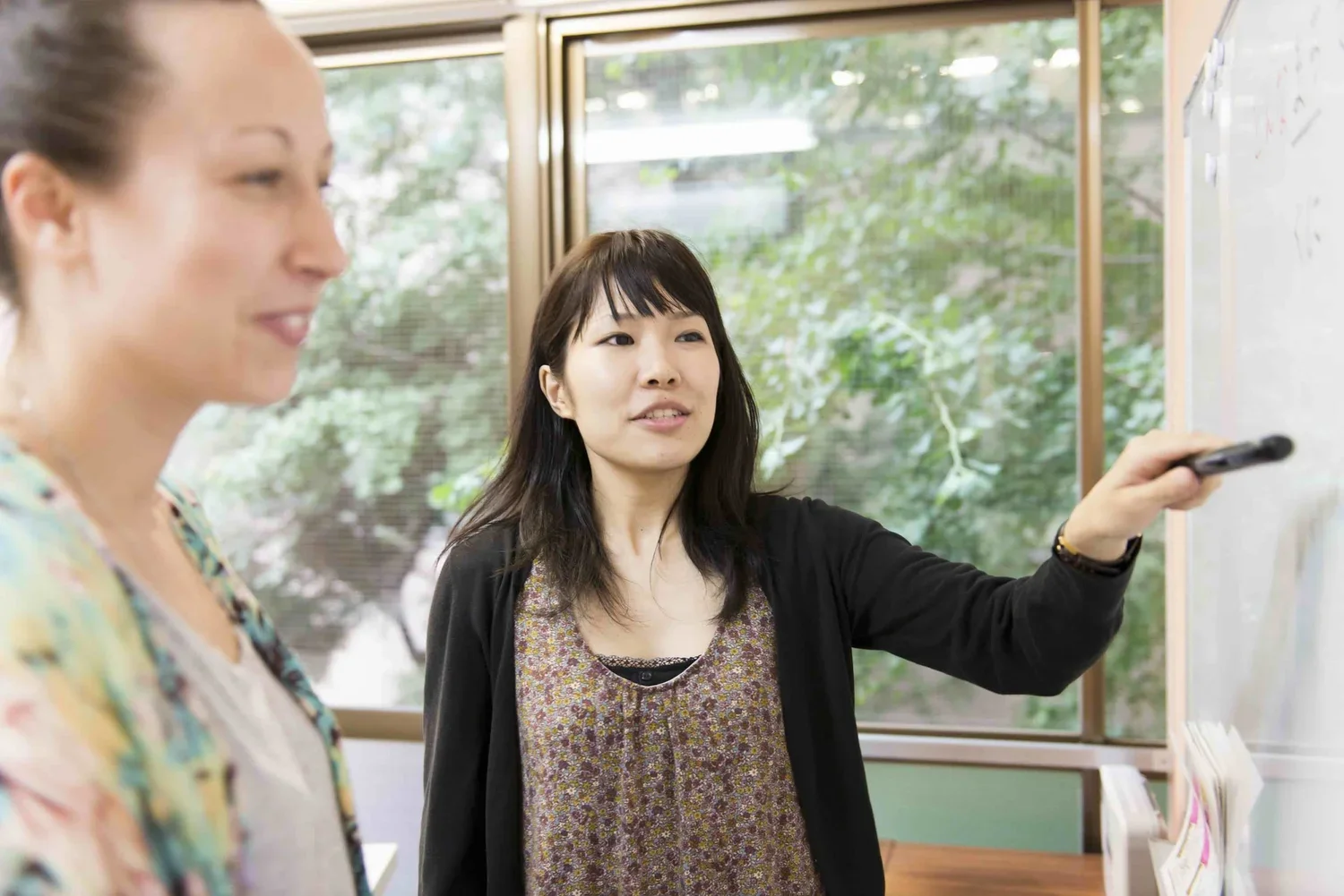 Studying Japanese. Two women in a sclassroom, one is pointing at a whiteboard while the other looks on. There are large windows with trees visible outside.