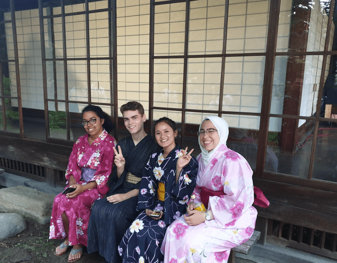 Japanese Language school in Kyoto. Four young students, three women and one man, sitting on a wooden bench outside a building with large window panes, dressed in colorful yukata and kimono, smiling and making peace signs.