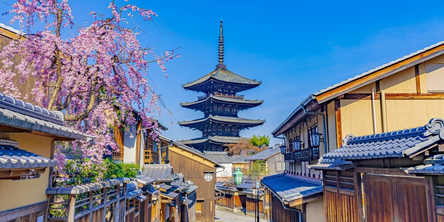 Traditional Japanese street with wooden buildings, pink cherry blossom trees, and a pagoda in the background under a clear blue sky.