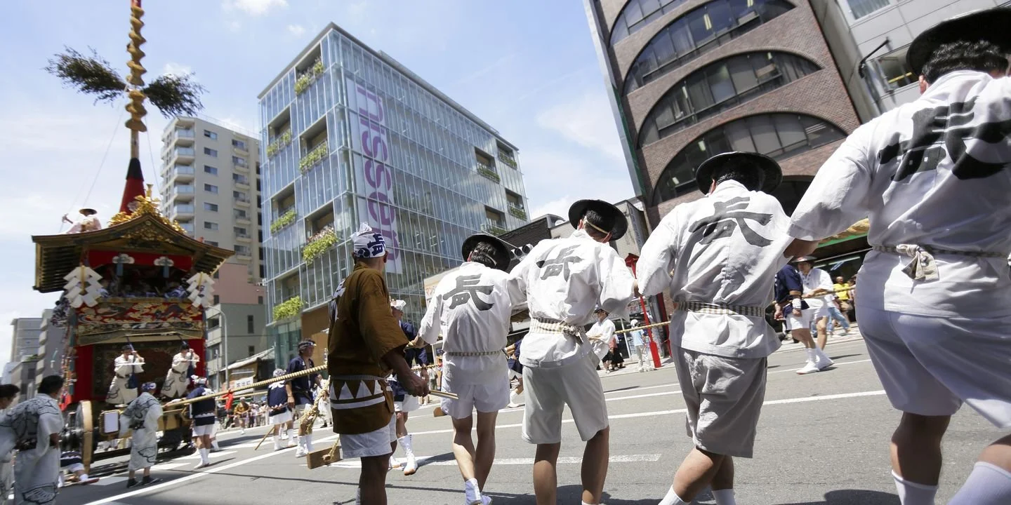 Group of people participating in a traditional Japanese festival parade, pulling a decorative float through city streets on a sunny day.