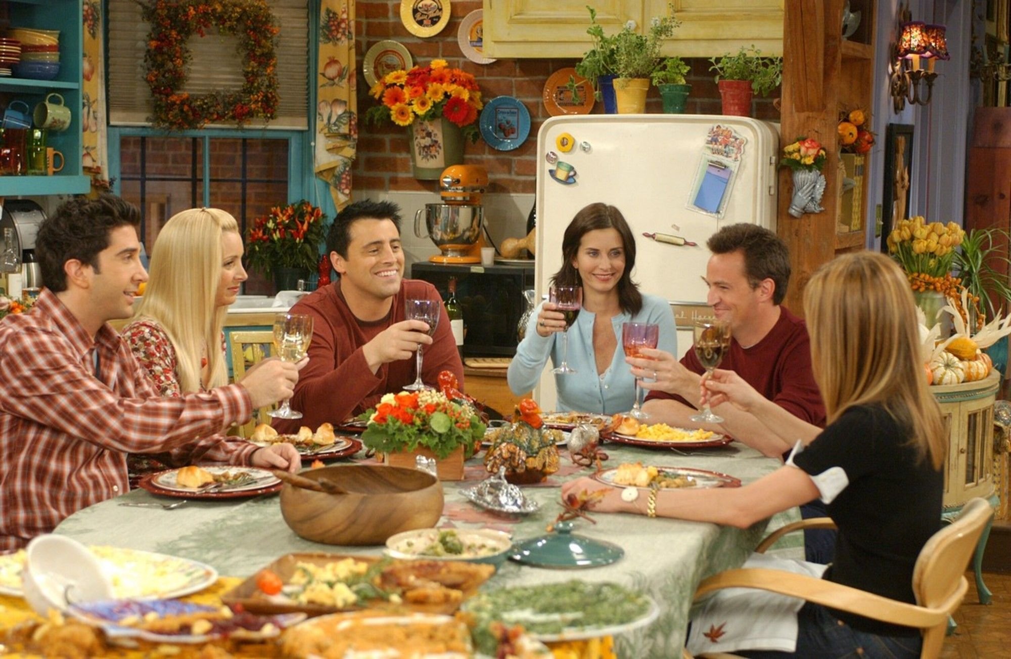 A scene from the TV show Friends, inspiring our Friendsgiving event at Petit and Keet. The cast gathers around Monica's kitchen table for Thanksgiving dinner, raising wine glasses in a toast, surrounded by fall decor.