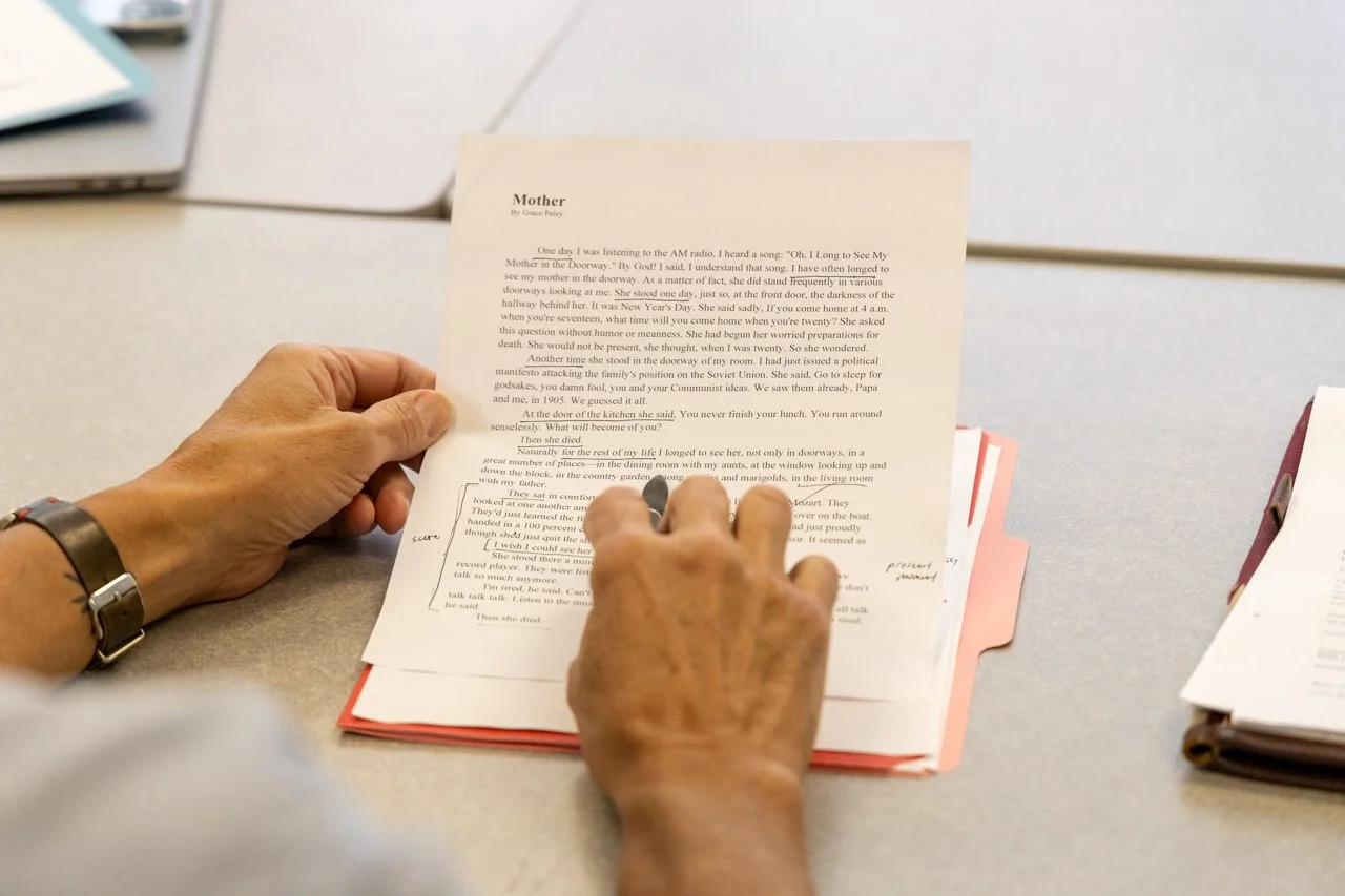 A person with a wristwatch is reading printed pages with handwritten notes and annotations on a table, with additional papers and files nearby.