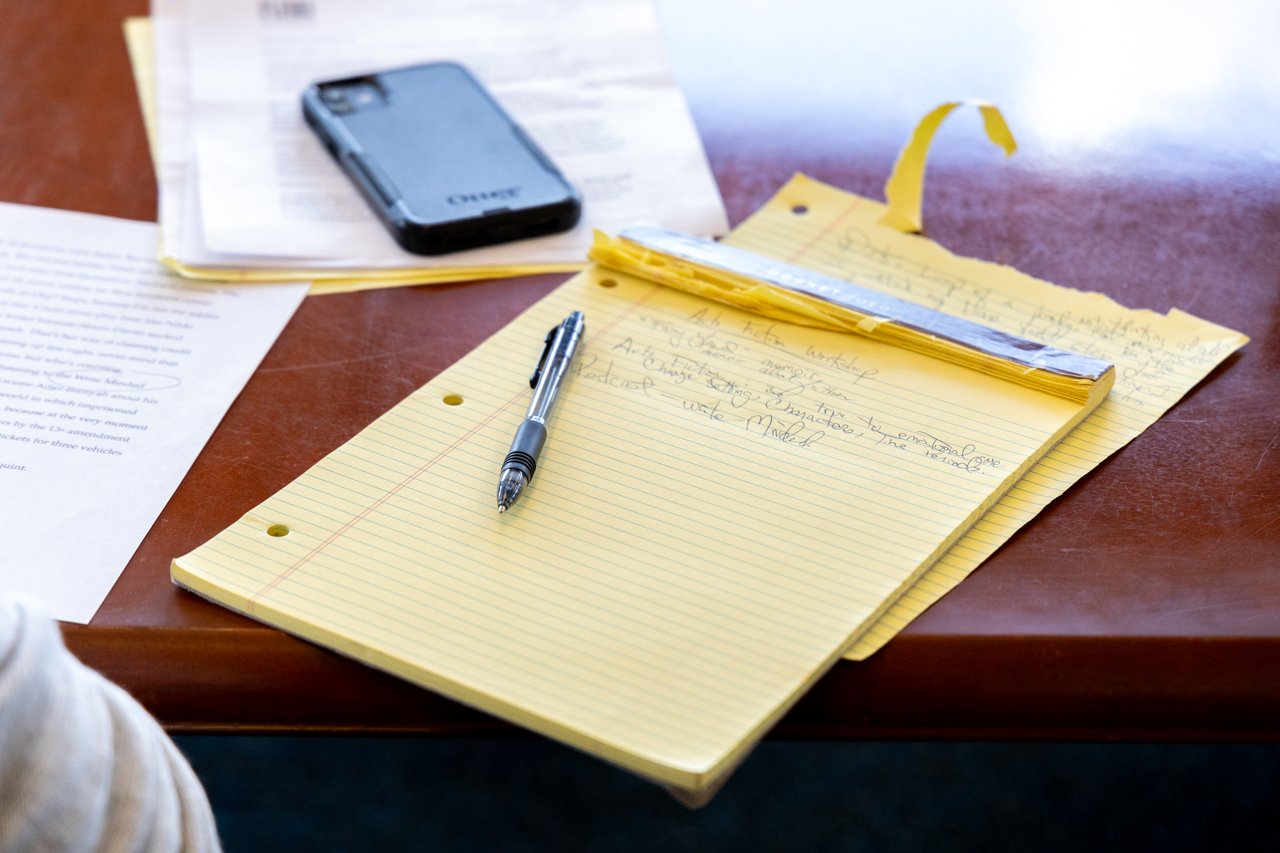 A yellow legal pad with handwritten notes and a black pen resting on it, a ruler on top, and a smartphone on papers on a wooden desk.