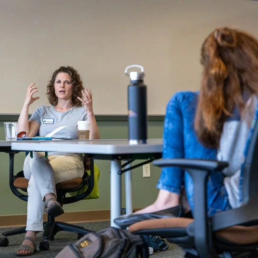 Two women sitting at a table in a room, one with curly hair and a gray t-shirt and the other with long hair and a blue jacket. The woman with curly hair appears to be talking or explaining something, with her hands raised. The table has a water glass, a disposable coffee cup, and a black water bottle.