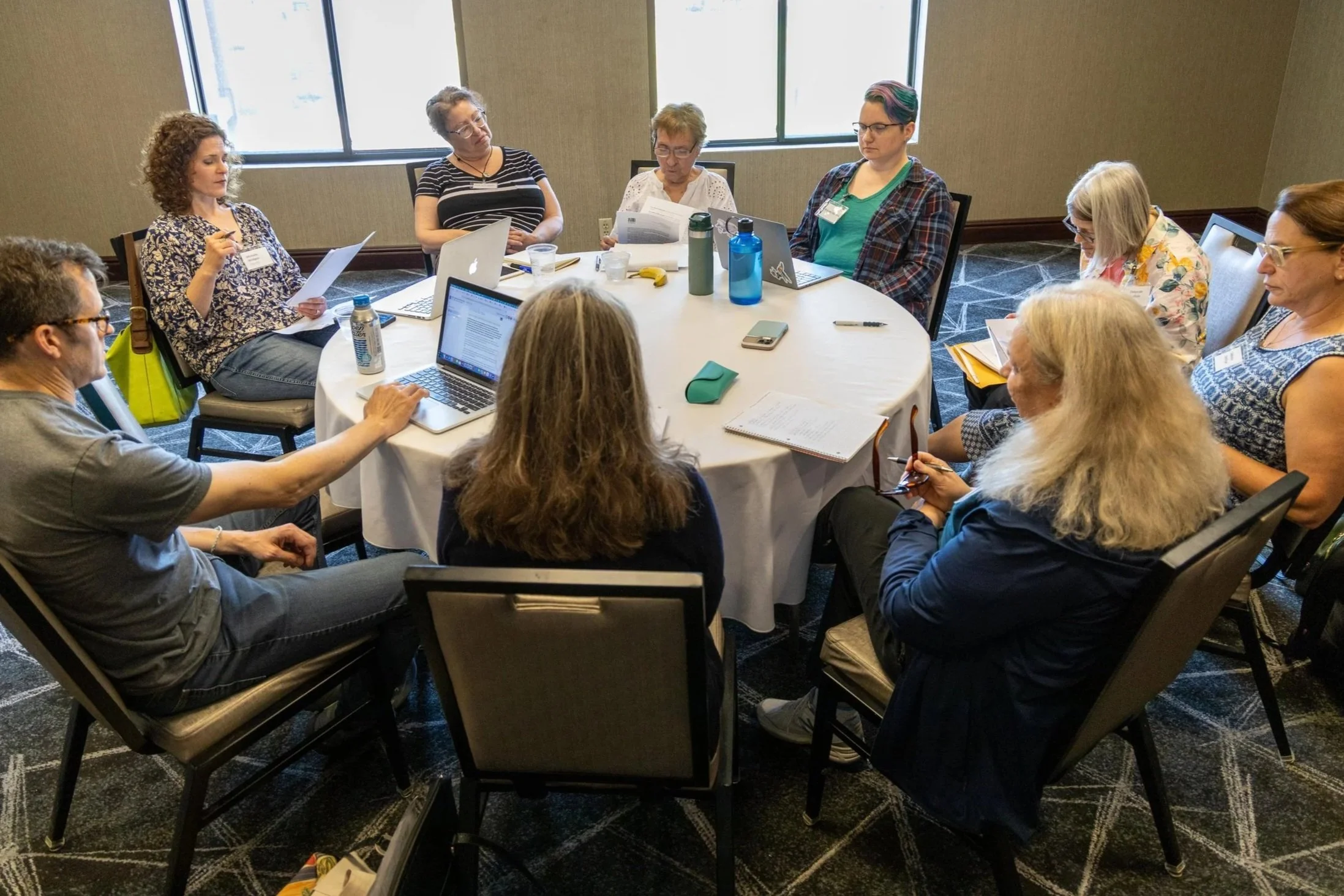 A group of women seated around a round table during a meeting, some with laptops, papers, and water bottles.
