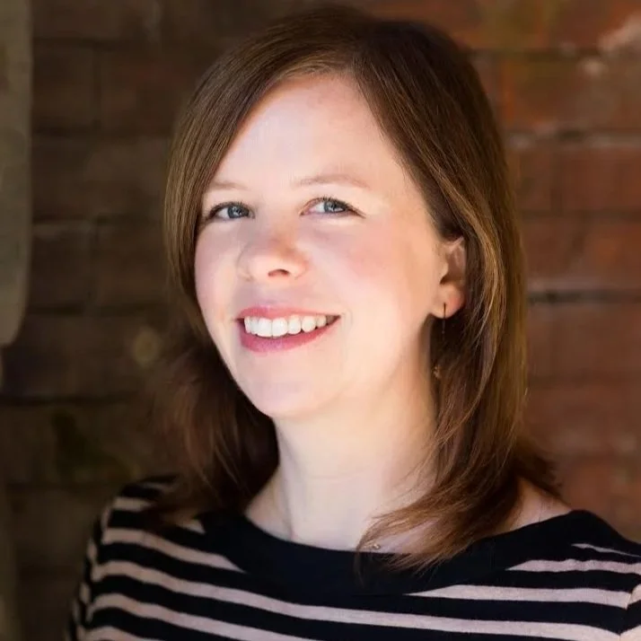 A woman with shoulder-length brown hair, blue eyes, and fair skin, smiling, wearing a black and white striped shirt, standing in front of a wooden background.