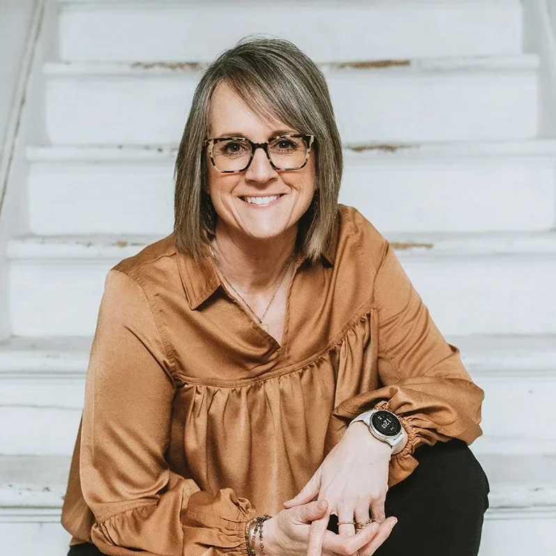 A middle-aged woman with shoulder-length light brown hair, wearing glasses with tortoiseshell frames, a brown satin blouse, and a smartwatch, sitting on white stairs and smiling at the camera.