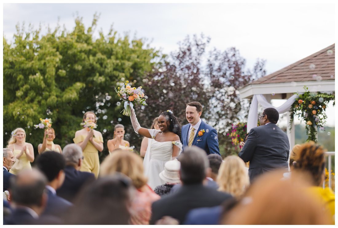 bride and groom walk down the aisle after ceremony and he lifts her bouquet in a cheer as bubbles surround them