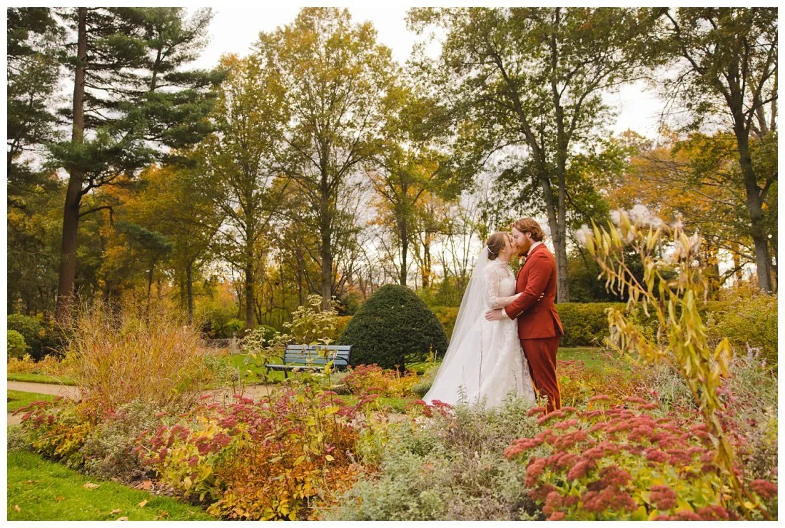 bride and groom kiss in autumn landscape with flowers in foreground and trees with fall foliage in background