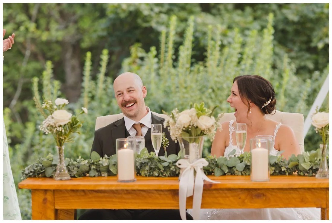 bride and groom big laughs during wedding toasts, with greenery and candles on the table in front of them