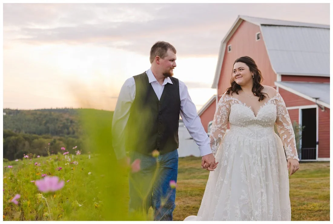 bride and groom hold hands and look at each other with red barn and mountains in the background at golden hour