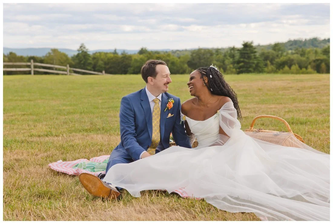 bride and groom laugh while sitting in open field with mountains in background