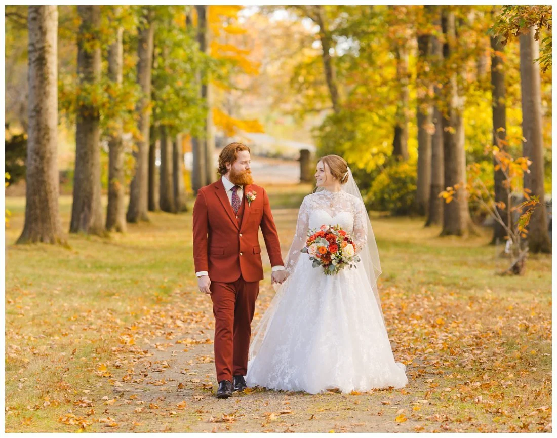 yellow fall leaves cover the tree lined pathway at glen magna farms, while the wedding couple holds hands and walks