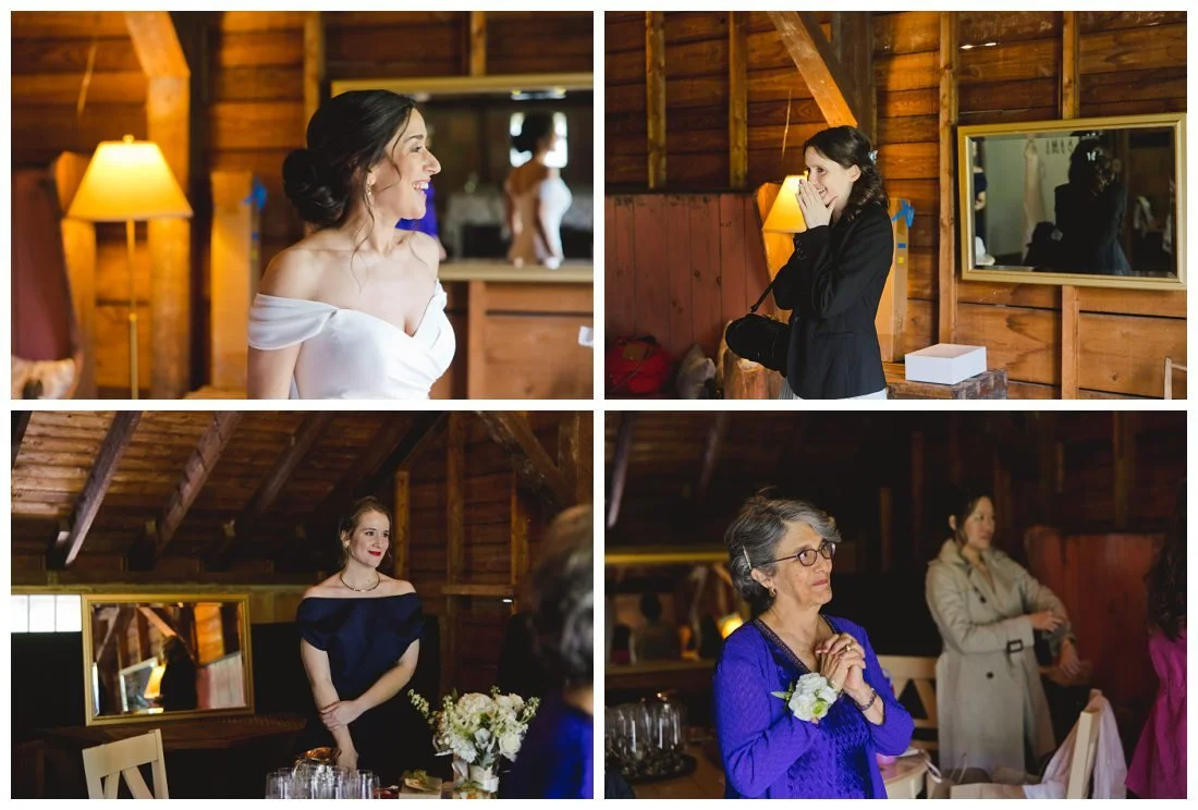 bride gets ready in a renovated barn and smiles while looking in the mirror and her mom and bridesmaids look on