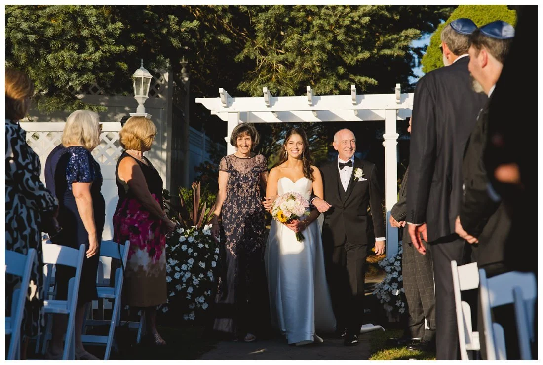 rachel, glowing in the sunlight, walks down the aisle with her parents