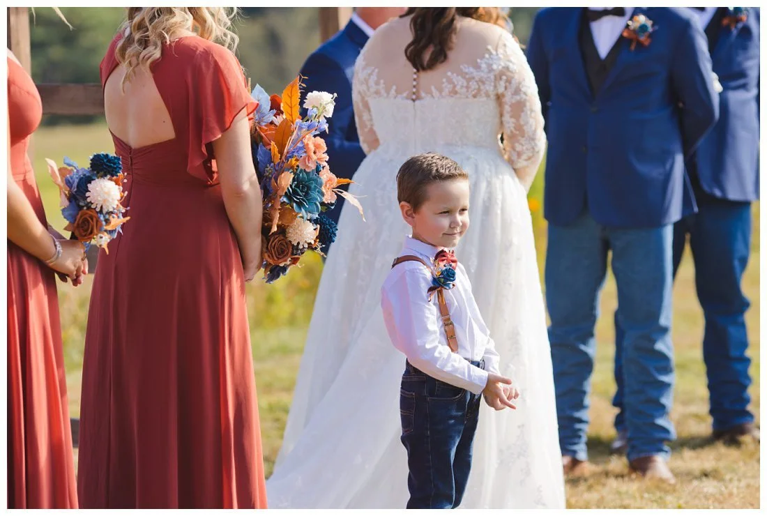 cute ring bearer with jeans and suspenders looks at guests during wedding ceremony