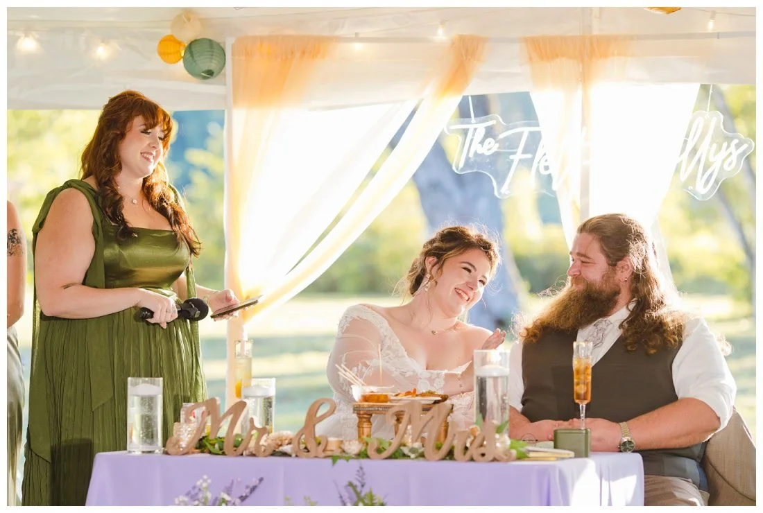 bride laughs and pats her groom on the arm as her sister gives toast