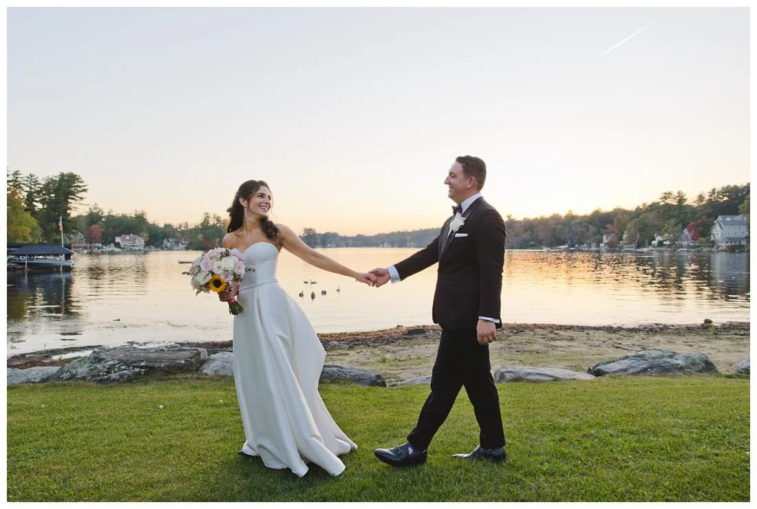 bride leads groom on lawn in front of of lake
