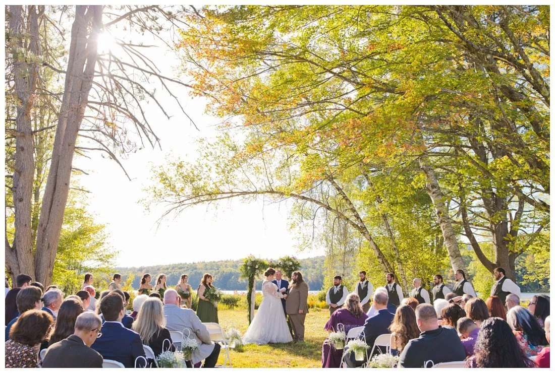 wide angle photo of wedding ceremony on lake winnipesaukee with trees all around them and lake behind them