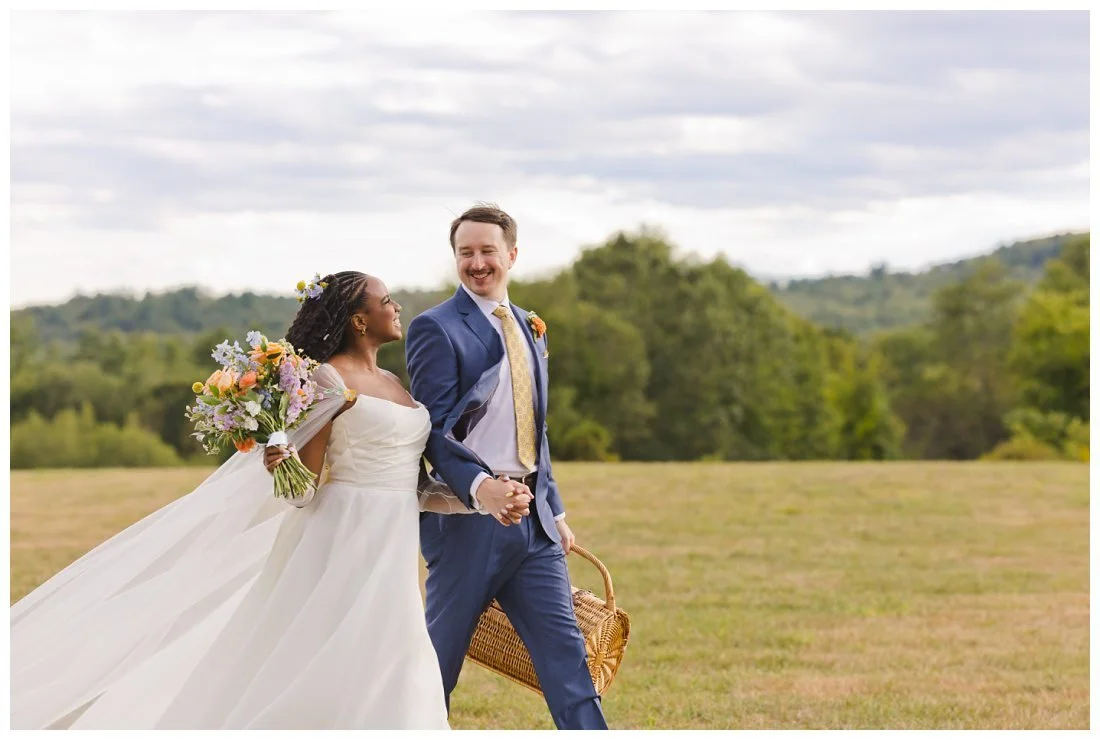 bride and groom walk in field looking at each other groom carries picnic basket
