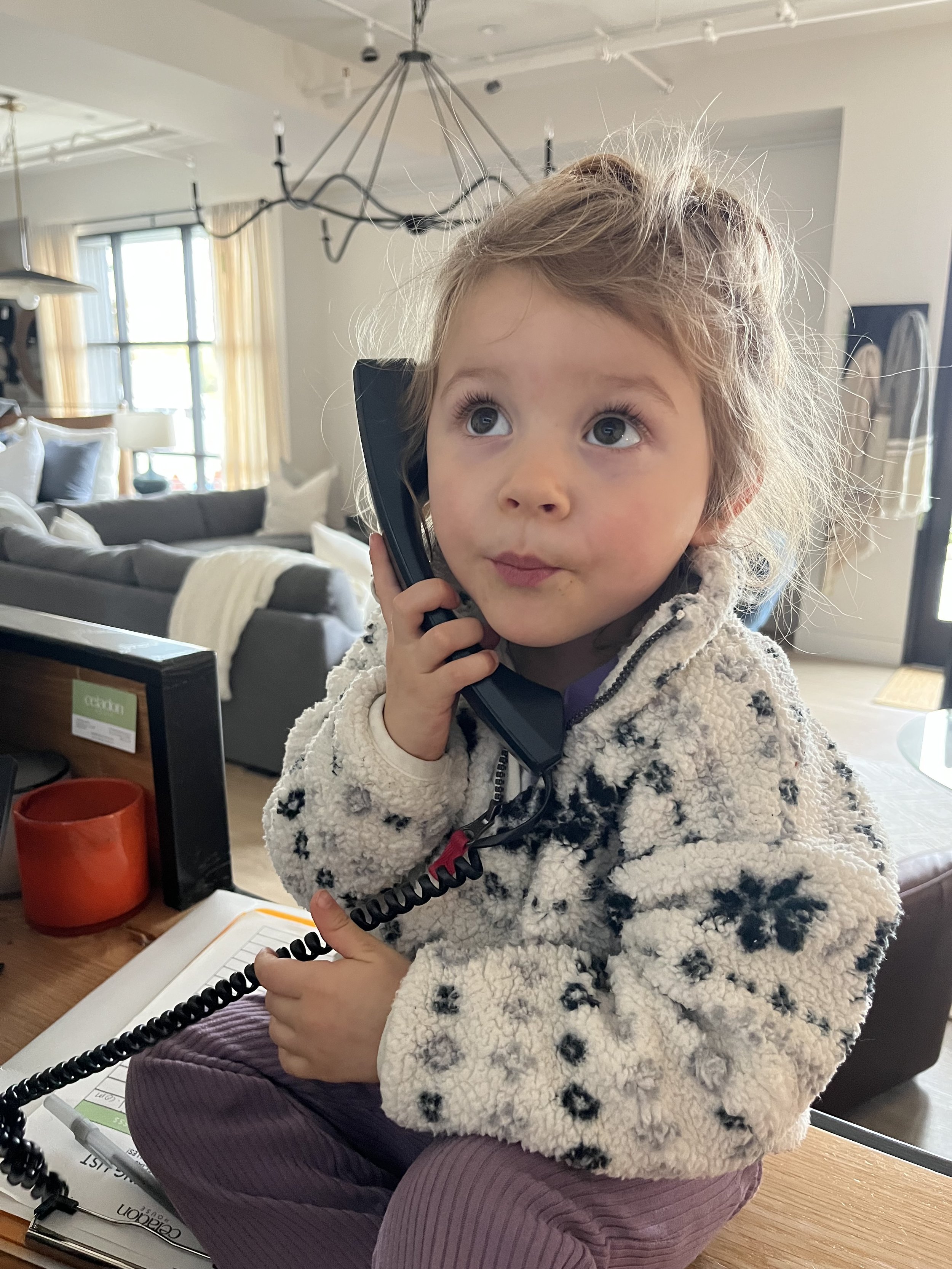 A young girl with curly blonde hair and big blue eyes, wearing a cream-colored jacket with black patterns, sitting at a table and talking on a corded telephone in a brightly lit living room.