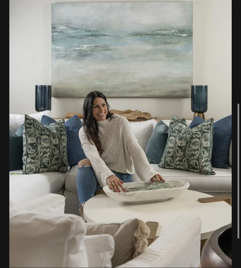 A woman with long dark hair, wearing a cream sweater, sitting on a white couch surrounded by patterned and solid blue pillows, smiling and touching a large decorative bowl on a white coffee table in a modern living room decorated with a large abstract ocean painting and blue table lamps.