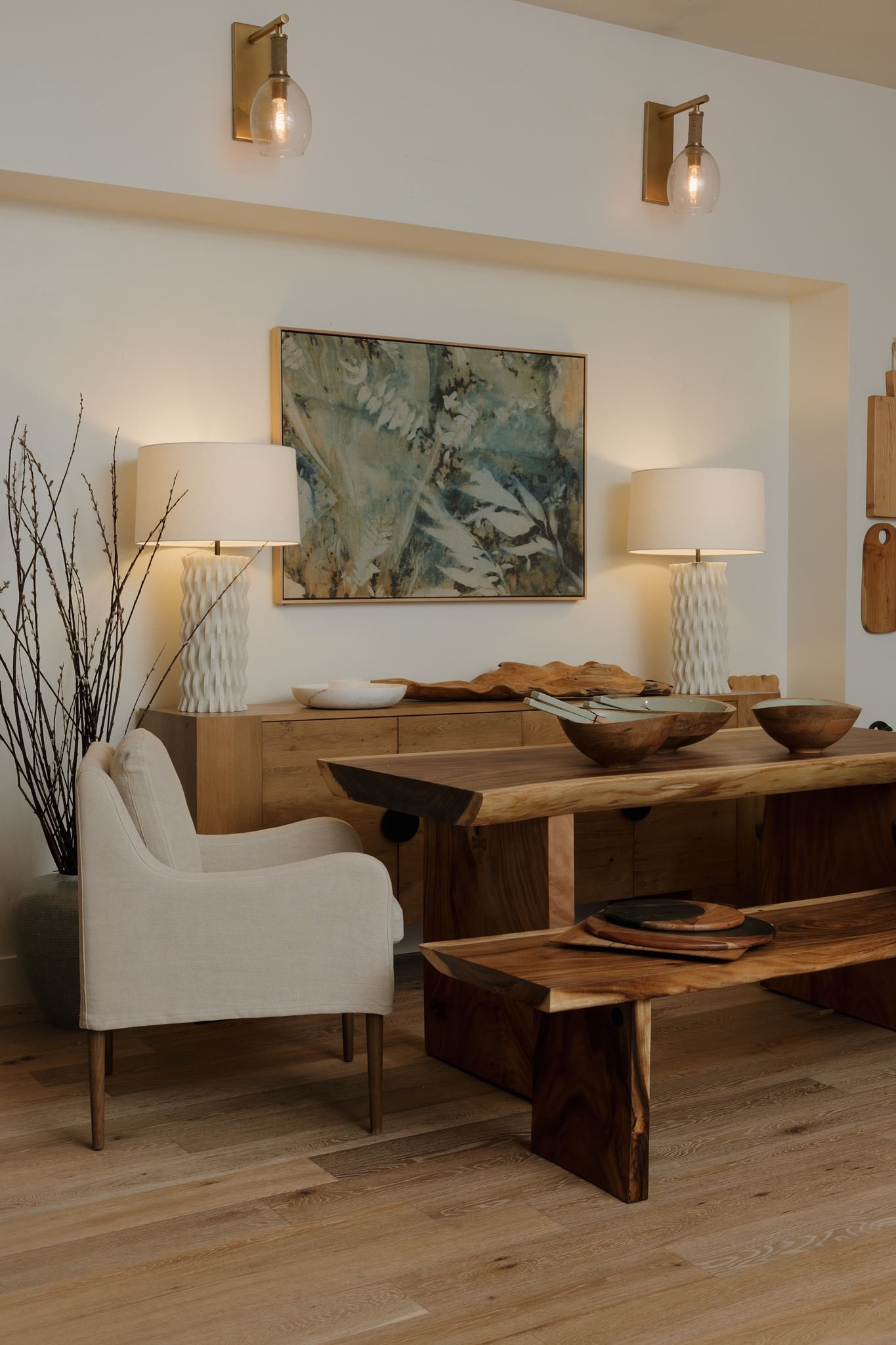 A living room corner with two brass wall-mounted light fixtures, a framed abstract painting, white ceramic lamps, a wooden sideboard, a cream armchair, and a rustic wooden dining table with bowls and plates.