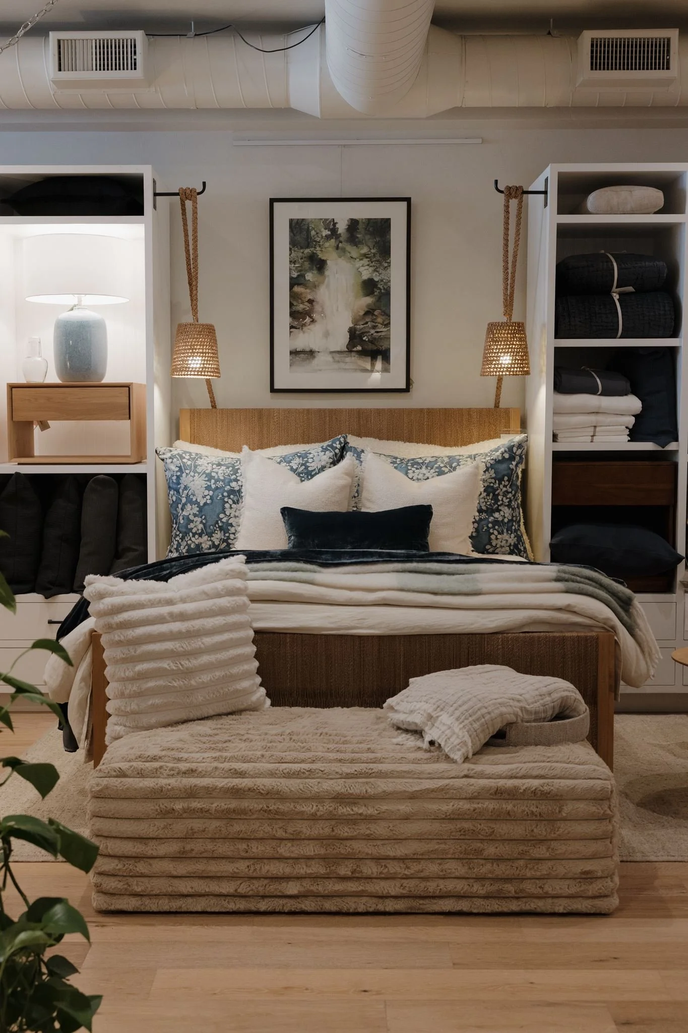 A cozy bedroom with a bed featuring a rattan headboard, decorative pillows, and layered blankets. Flanking the bed are hanging woven lamps. Behind the bed is a framed landscape painting. To the left is a white shelving unit with a blue vase and a table lamp, while on the right is a similar shelving unit with folded blankets and boxes. The room has light wood flooring and above, large white ductwork is visible on the ceiling.