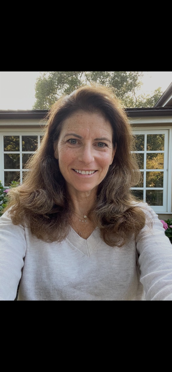 A woman with long, wavy brown hair smiling outdoors in front of a house with a large window and trees in the background.