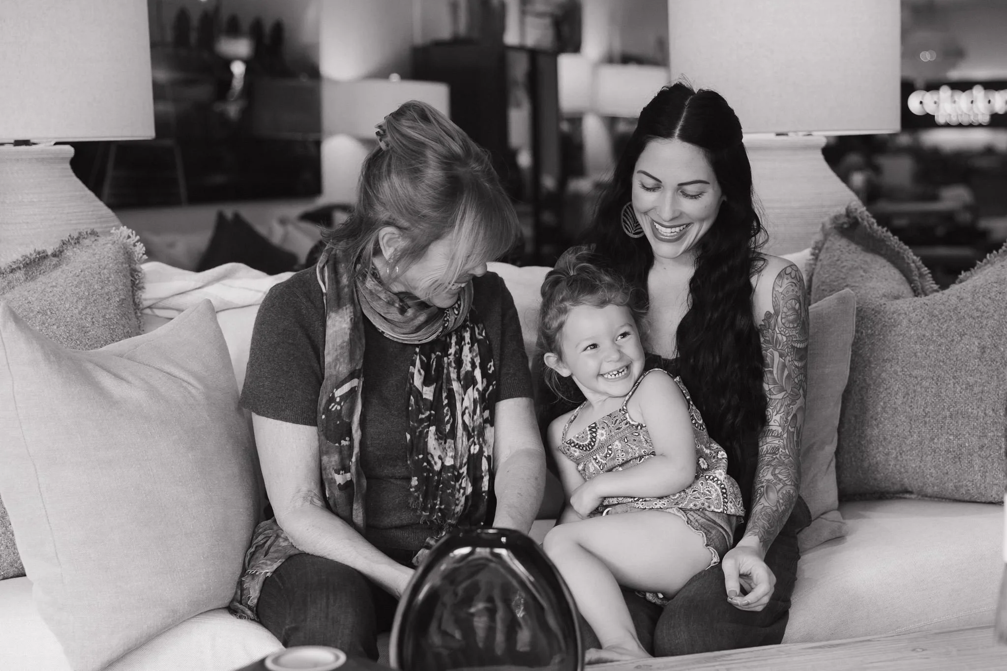 Black and white photo of three women and a young girl sitting on a couch, looking at something in front of them, smiling and enjoying a moment together in a living room.