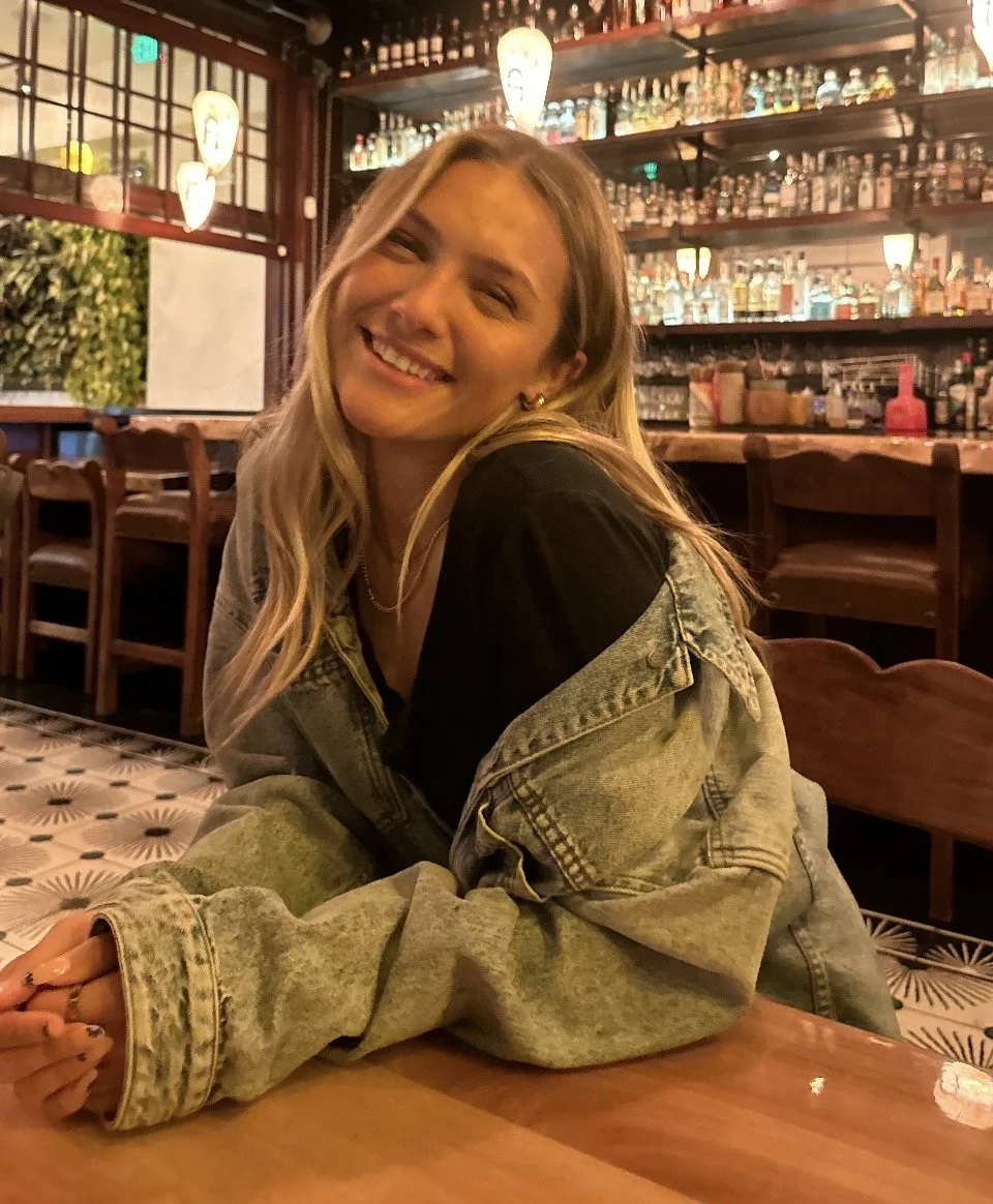 A young woman smiling and leaning on a wooden table inside a bar with a well-stocked back bar and decorative lighting.