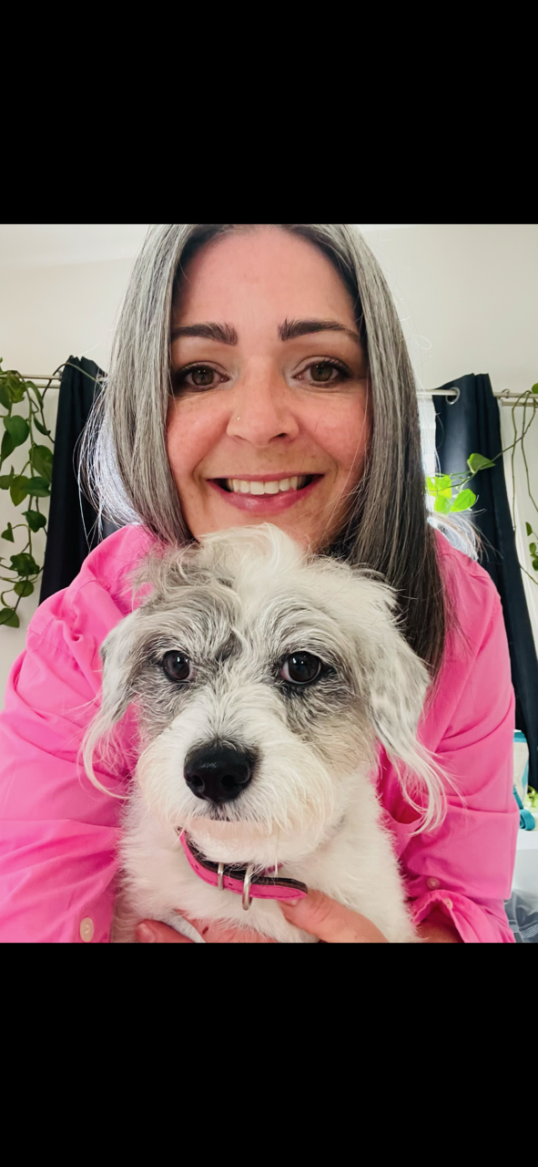 A woman with shoulder-length gray hair and a pink jacket holding a small, white and gray dog with a pink collar indoors, with green plants and dark curtains in the background.