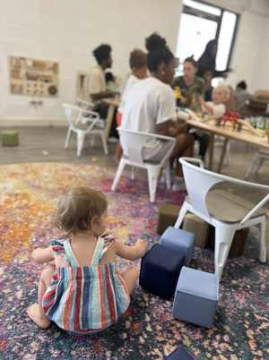 A young child sitting on a colorful rug on the floor, playing with blue and gray foam blocks, in a lively indoor space with a group of people gathered at a table in the background.