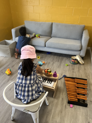 A young girl sitting on a chair playing a small toy piano, with a pink hat and a plaid dress. A boy is kneeling on the floor near a sofa, playing with toys on the cushions. There are various toys scattered on the floor, including a xylophone.