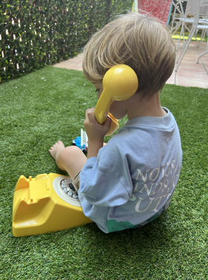 A young child with blonde hair playing with a yellow toy telephone on a green artificial grass surface outdoors.