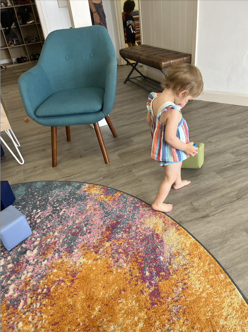 A young girl with short hair wearing a striped sleeveless dress stands barefoot on a wooden floor, holding a small object near a green box, in a room with a colorful round rug and a teal armchair.