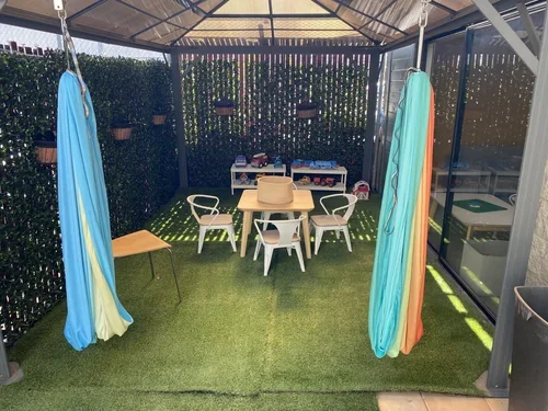 Outdoor patio with artificial grass, white chairs and table, hanging colorful fabric dividers, potted plants on the hedge, outdoor furniture, and a trash bin.