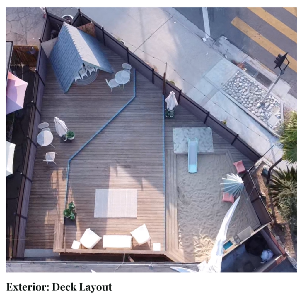 An aerial view of a rooftop deck with wooden flooring, outdoor furniture including a round table with chairs, a small slide, an umbrella, a lounge area with cushions, and potted plants. The deck is enclosed by fencing and overlooks a sidewalk, street, and crosswalk.