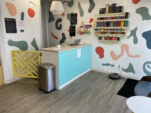 Interior of a colorful shop with abstract wall art, a small counter with a laptop, a trash can, and a robot vacuum on the floor. Shelves on the wall hold containers of various colored items, possibly supplies or ingredients.