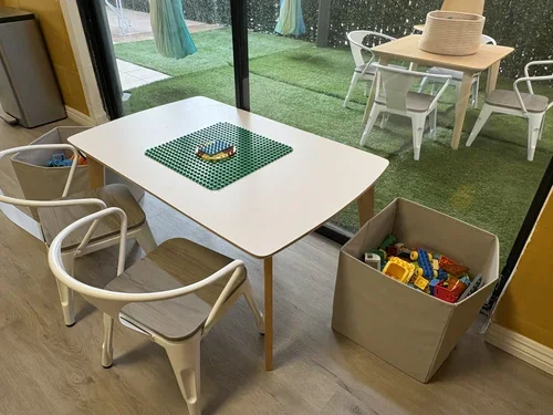 Child's play area with a white table, three white chairs, a box of colorful plastic toy building blocks, and a green Lego base plate with a bracelet on top, next to a large window showing an outdoor patio with a table and chairs and a grassy yard.