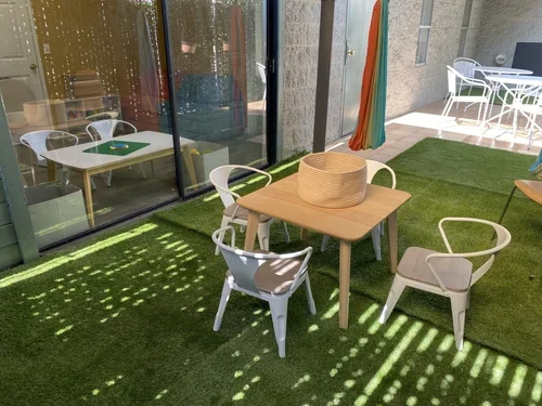Outdoor patio area with a wooden table, four white chairs, a large woven basket on the table, and an artificial grass surface. Behind a glass sliding door, an indoor dining table with white chairs and a toy on the table are visible.